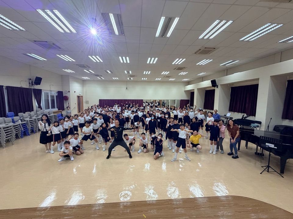 一大群穿校服的小學生在禮堂裏擺出運動姿勢,前方一位成年人雙腳張開站立。