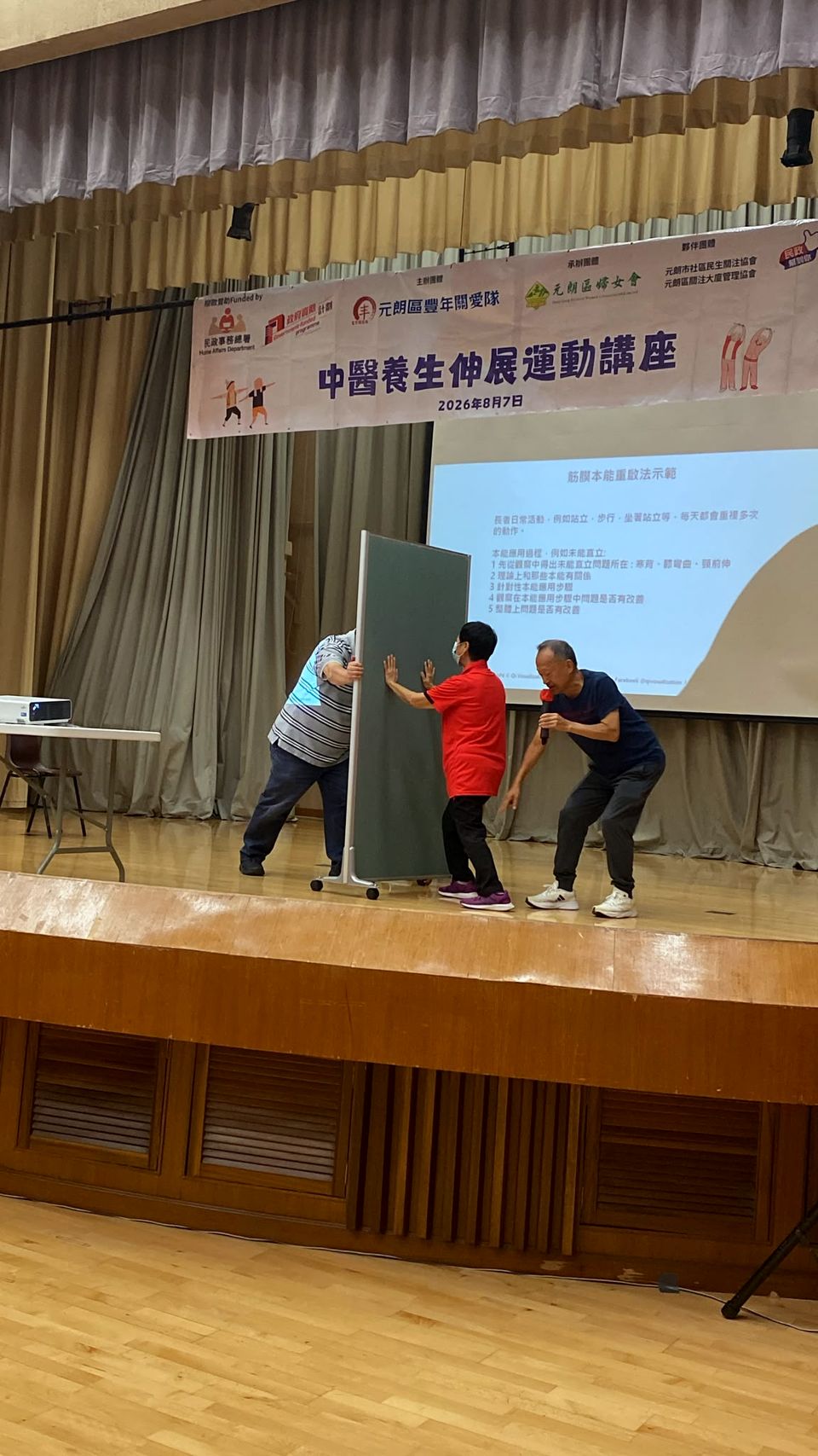 舞台上，幾個人雙手撐著一塊直立的板，身體前傾，做伸展運動。