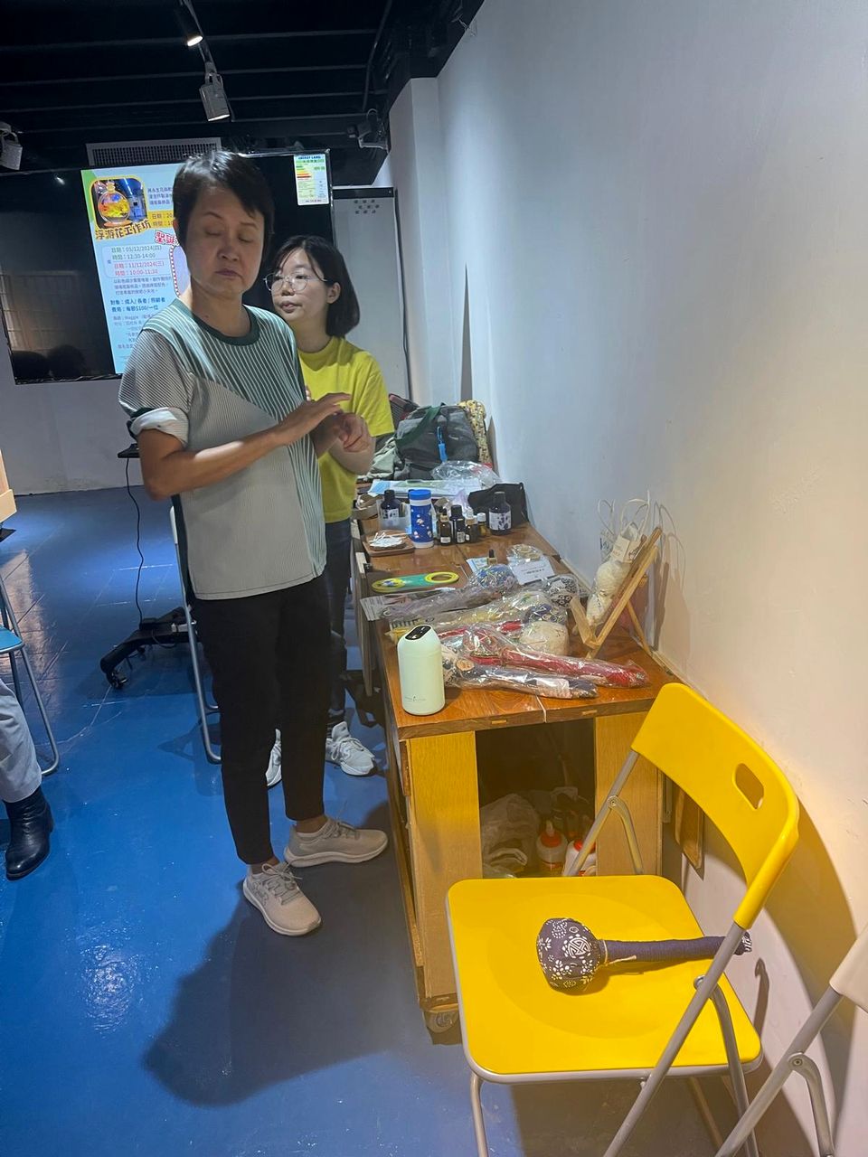 兩位女士站在擺放了手工材料的桌子旁，她們前面的黃色椅子上放著一件手作成品。