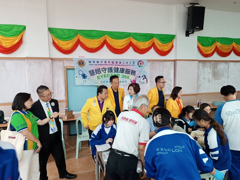 穿黃色外套的成年人和穿學校運動服的學生，聚在一張桌子旁，上方掛著寫有EyeCare 365的橫額。