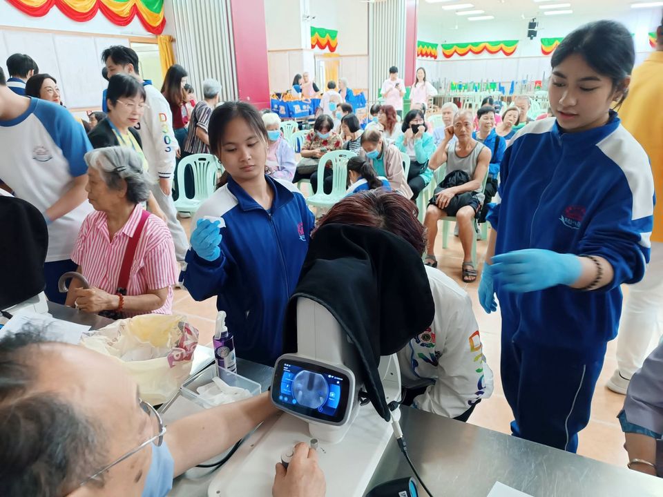 在熱鬧的禮堂裏，兩位穿學校運動服的學生義工站在眼底照相機旁，其中一位拿著一張新的下巴托墊紙；一位男士正為坐著的訪客操作照相機。