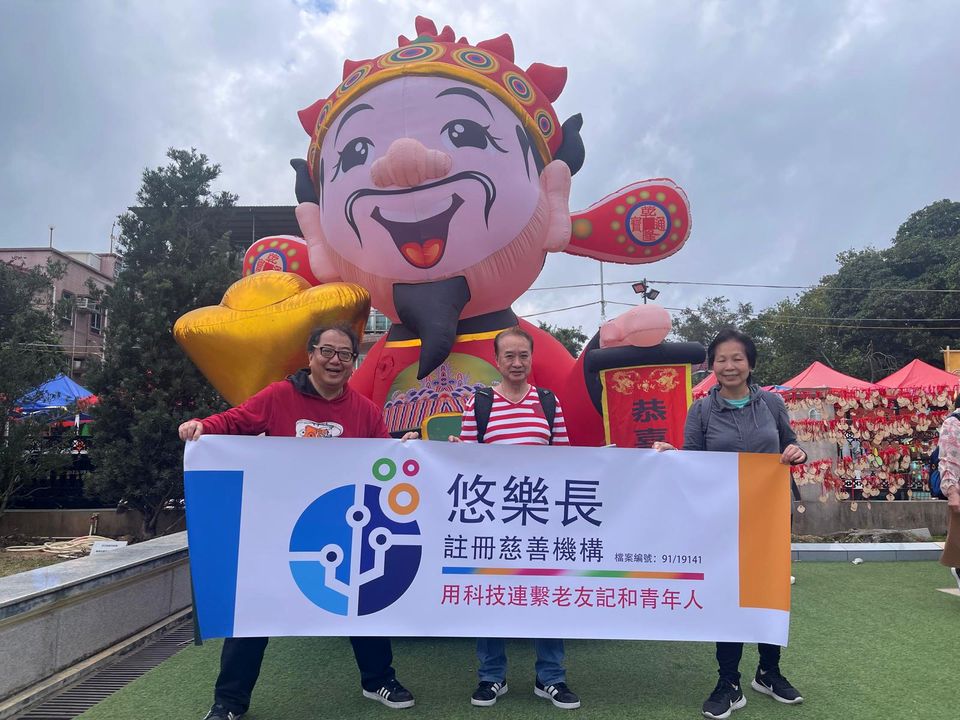 三個人在草地上手持悠樂長的橫額，身後是一個巨型的農曆新年充氣擺設。