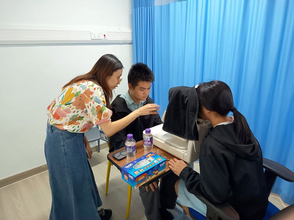 一位導師俯身在兩名穿校服的學生旁邊，其中一名學生正望進眼底照相機。