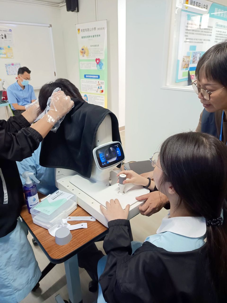 一名女學生操作眼底照相機，導師站在她身旁；一位同學坐在下巴托架前，頭上蓋著一塊深色的布。